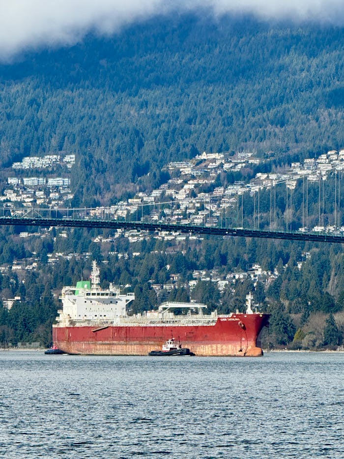 A cargo ship sails under a bridge in Vancouver, framed by lush forests and cityscape.